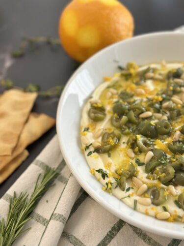 A bowl of creamy dip topped with green olives, pine nuts, and herbs, garnished with orange zest, accompanied by pita bread and an orange in the background.