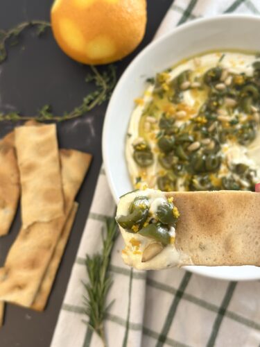 A close-up of a hand holding a piece of pita bread topped with a spread that includes green olives and spices, with a bowl of the spread in the background and an orange, herbs, and pieces of pita on the side.