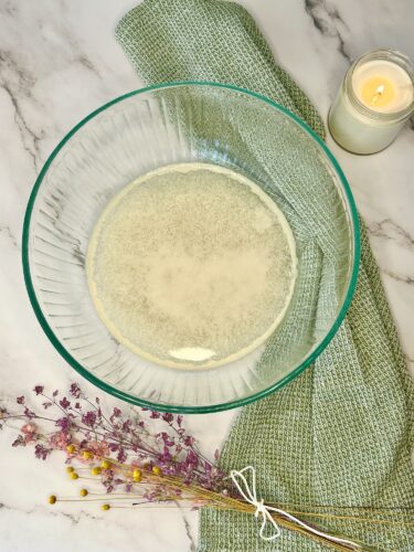 A glass bowl containing liquid, placed on a marble countertop with a green textured cloth, dried flowers, and a lit candle in the background.