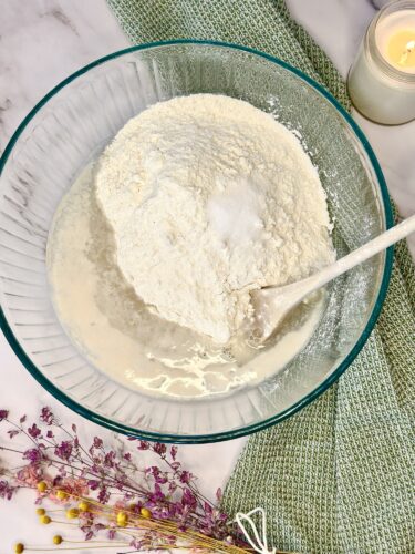 A glass bowl filled with a mixture of flour and liquid, with a wooden spoon resting inside, arranged on a textured green cloth, with dried flowers and a candle in the background.