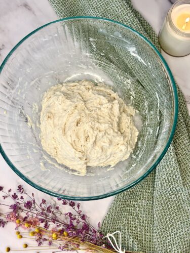 A glass bowl containing a thick, unrisen dough mixture, placed on a textured green cloth with dried flowers and a lit candle in the background.