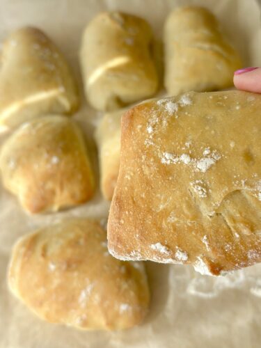 A close-up of a hand holding a piece of freshly baked bread, with several loaves in the background.