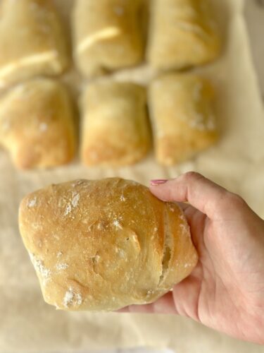 A hand holding a freshly baked bread roll, with several more rolls in the background on parchment paper.