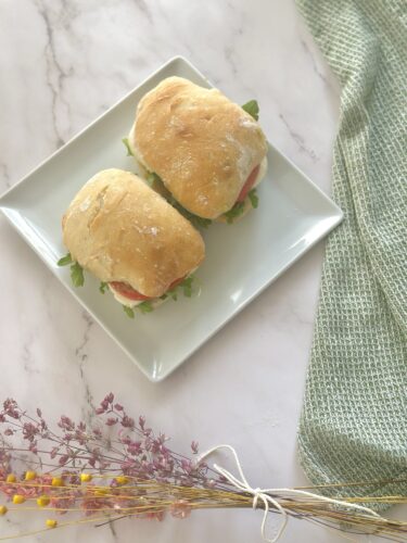 Two small sandwiches with fresh greens and slices of meat, served on a square white plate, with a textured green cloth and dried flowers in the background.