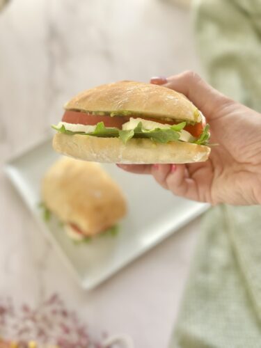 A person holding a sandwich filled with lettuce, tomato, and cheese, with another sandwich in the background on a plate.