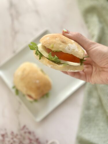 A hand holding a sandwich with lettuce, tomato, and onion, with another sandwich on a plate in the background.