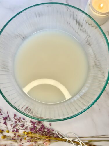 A clear glass bowl filled with a light-colored liquid, surrounded by dried flowers and a softly glowing candle in the background.