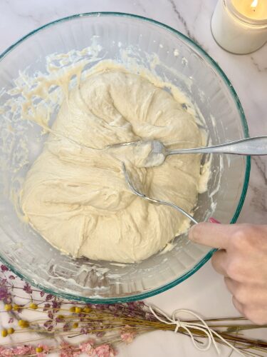 A close-up of a bowl of dough being mixed with two forks, surrounded by dried flowers and a candle.