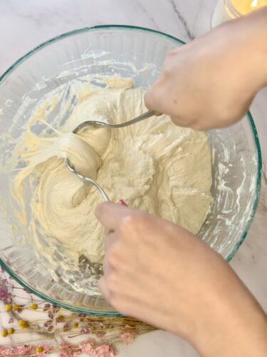 Hands mixing dough in a glass bowl with a spatula, surrounded by decorative flowers.