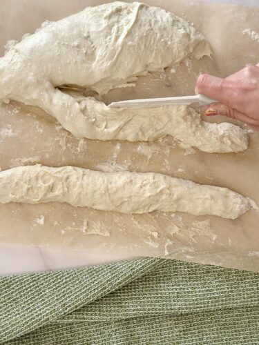 A hand using a knife to cut dough on a floured surface, with elongated pieces of dough and a green textured cloth in the foreground.