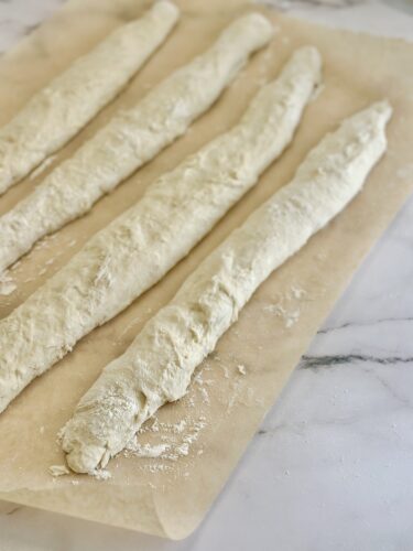 Four elongated pieces of raw dough resting on parchment paper, sprinkled with flour.