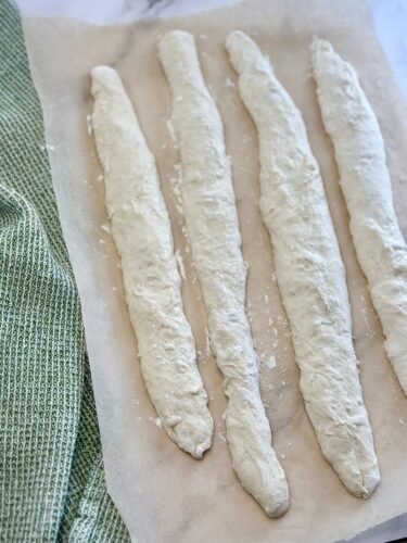 Four elongated pieces of uncooked bread dough on parchment paper, with a green kitchen towel partially visible in the background.