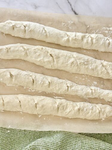 Raw dough shaped into long strands resting on parchment paper, with a cloth underneath.
