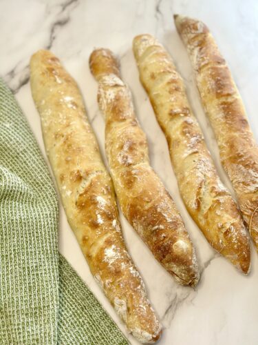 Four freshly baked baguettes arranged on a marble countertop with a green kitchen towel beside them.