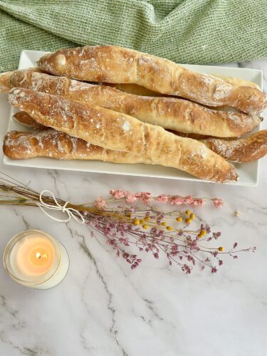 A platter of freshly baked, elongated bread sticks dusted with flour, accompanied by a lit candle and dried flowers on a decorative table setting.