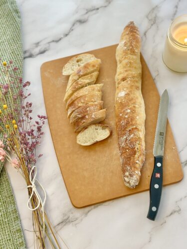 A freshly baked baguette partially sliced on a wooden cutting board, accompanied by a knife and decorative dried flowers.