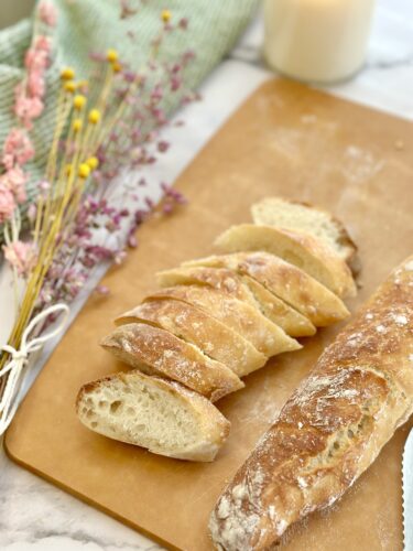 Sliced fresh bread on a wooden cutting board, next to a whole baguette and dried flowers in the background.
