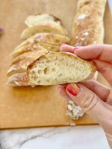 A close-up of a hand holding a slice of freshly baked bread, with a loaf and sliced portions in the background on a wooden cutting board.
