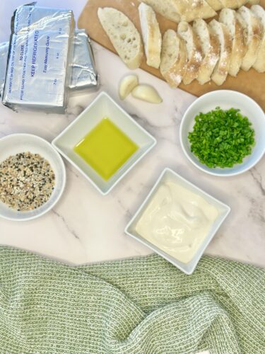 Ingredients arranged on a marble surface, including sliced bread, blocks of cream cheese, minced garlic, chopped green onions, olive oil, and a bowl of mixed seasonings, accompanied by a textured green cloth.