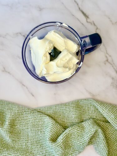 A blender filled with creamy white ingredients, possibly for a dip or spread, sitting on a marble countertop with a green patterned kitchen towel nearby.