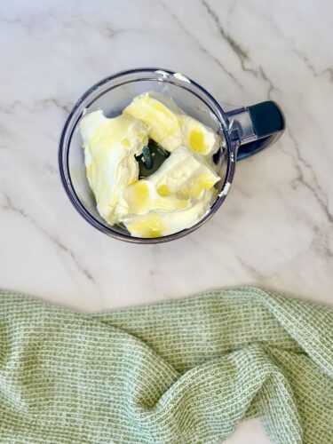 A clear mixing bowl containing a mixture of cream or yogurt, topped with green herbs and drizzled with olive oil, placed on a marble countertop next to a green textured kitchen towel.