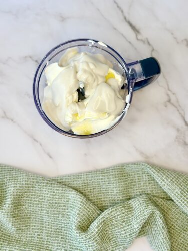 A clear blender container filled with a mixture of yogurt and yellow ingredients, placed on a marble countertop, with a green kitchen towel in the foreground.
