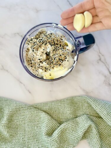 A top view of a mixing cup containing a creamy mixture topped with spices and ingredients, with garlic cloves held above it, and a green cloth underneath.