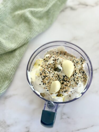 A close-up view of a food processor containing a mixture of cream, diced ingredients, and seasoning. There's a green kitchen towel in the background.