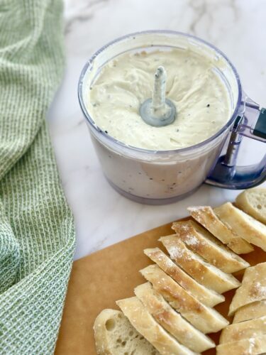 A food processor containing creamy dip with herbs, placed next to slices of bread on a wooden cutting board, with a green kitchen towel in the background.