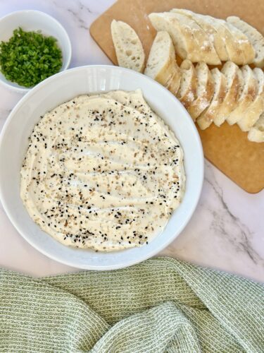 A bowl of creamy dip topped with black pepper, accompanied by sliced bread and chopped green herbs on a wooden cutting board.