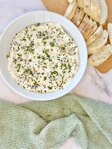 A bowl of creamy dip topped with chopped chives and sesame seeds, surrounded by sliced bread on a cutting board and a green kitchen towel.