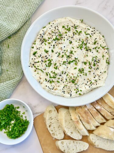 A bowl of creamy dip topped with chopped chives and black sesame seeds, accompanied by slices of crusty bread on a cutting board and a small dish of chopped chives on the side.