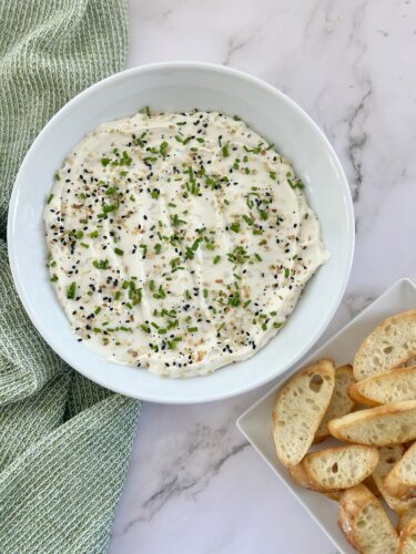 A bowl of creamy dip topped with chopped green onions and black sesame seeds, served with crispy toasted bread slices on the side.