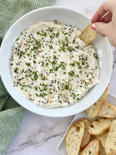 A bowl of creamy dip topped with chopped chives and black sesame seeds, with a piece of bread being dipped into it. Slices of toasted bread are arranged on a nearby plate.