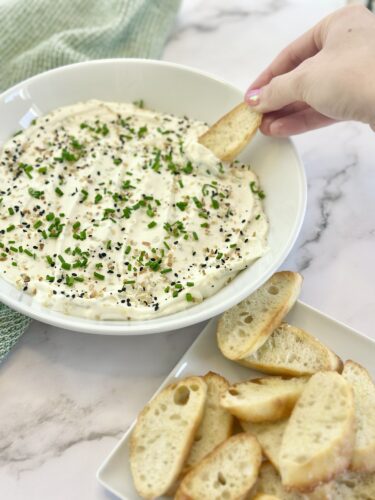 A hand dipping a slice of toasted bread into a creamy dip topped with chives and black pepper, served in a white bowl with additional bread slices on the side.