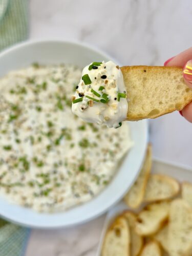 A hand holding a toasted bread slice topped with creamy dip, with a bowl of the same dip garnished with chives in the background.