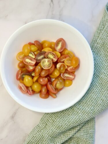 A white bowl filled with a variety of halved cherry tomatoes in different shades of red and yellow, placed on a marble surface next to a green textured towel.