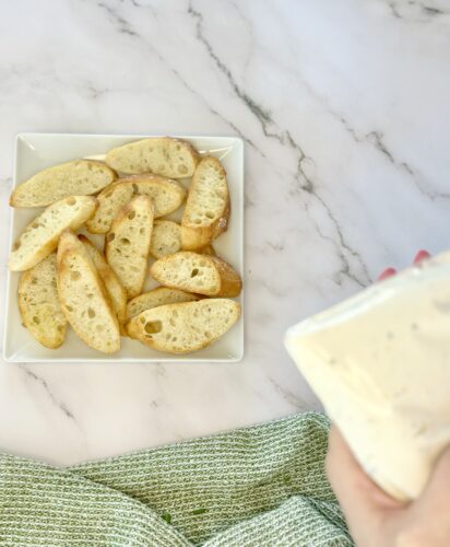 A hand holding a packet of sauce above a plate of toasted bread slices on a marble surface.