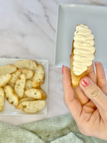 A hand holding a breadstick topped with creamy spread, with a plate of cut breadsticks in the background.