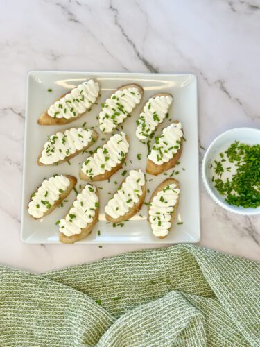 A platter of small toasted bread slices topped with creamy filling and sprinkled with chopped chives, accompanied by a small bowl of additional chopped chives.