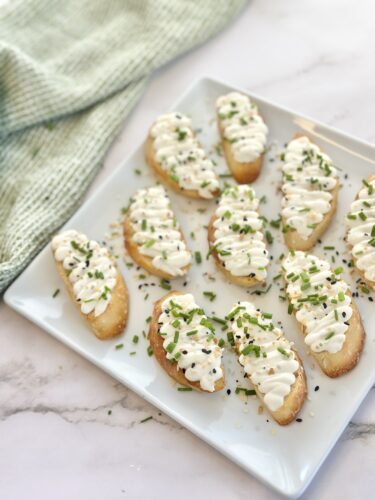 A platter of crispy bread slices topped with creamy white sauce and garnished with chopped chives and sesame seeds.