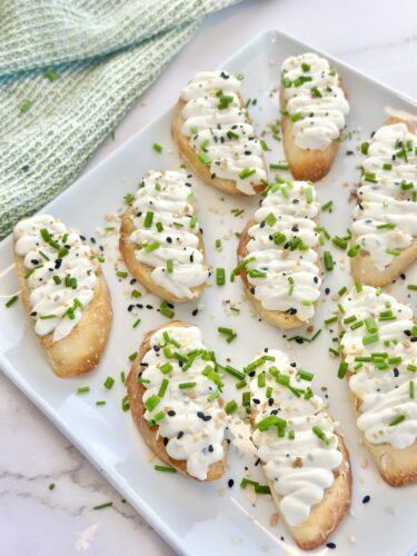 A platter of bite-sized pastry shells topped with creamy white frosting and sprinkled with chopped green onions and black sesame seeds.