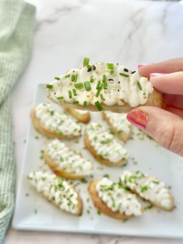 A hand holding a potato skin topped with sour cream and chopped chives, with more potato skins arranged on a plate in the background.