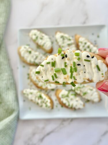 A hand holding a slice of bread topped with cream cheese and chopped chives, with more similar slices arranged on a plate in the background.