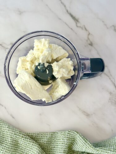 Top view of a food processor containing blocks of cream cheese on a marble countertop.