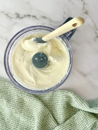 A top-down view of a food processor containing creamy, blended mixture, with a wooden spoon resting inside and a green kitchen towel peeking out below.