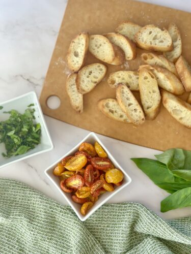 Sliced toasted bread arranged on a wooden cutting board, with a bowl of roasted cherry tomatoes and a dish of chopped fresh herbs beside it, surrounded by a light green cloth.