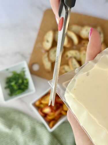 A person using scissors to cut open a packet of cream cheese, with baked potato slices and diced vegetables visible in the background.