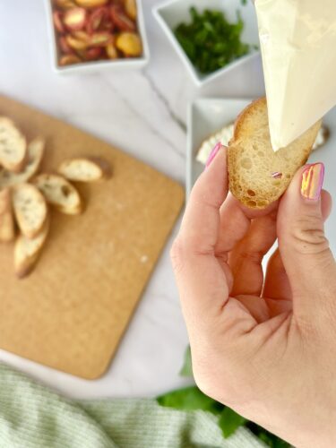 A hand holding a piece of toasted bread while piping cream onto it, with chopped herbs and sliced fruits in the background on a wooden cutting board.