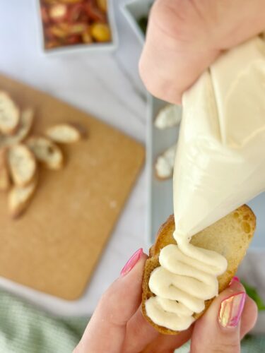 A hand holding a pastry bag is seen piping creamy frosting onto a small piece of bread, while additional bread slices are visible on a wooden cutting board in the background.